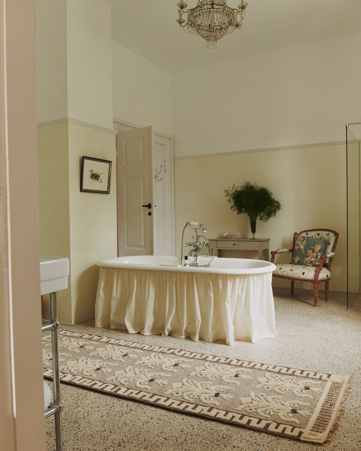 A inspiaring bathroom with a handmade, floral patterned rug, set in a classic bathroom with beige walls, a chandelier, and a floral armchair.