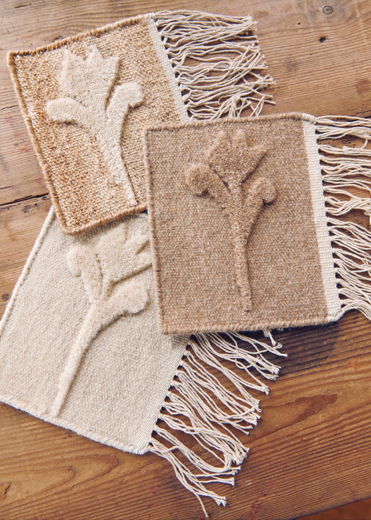 Three woven fringed rug samples in natural beige and cream tones, each featuring a raised botanical flower design, arranged on a rustic wooden floor.