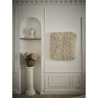 A square shag tapestry in a warm cream and beige tone hangs on a white paneled wall in a styled interior setting. 